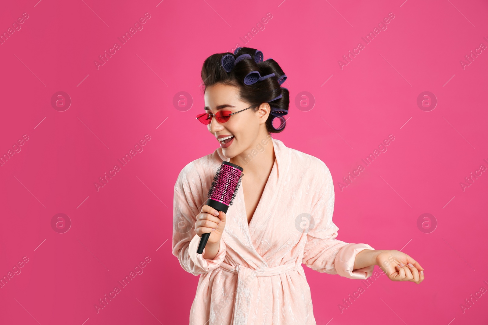 Photo of Happy young woman in bathrobe with hair curlers and sunglasses singing into hairbrush on pink background