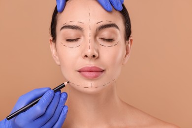 Doctor drawing marks on woman's face for cosmetic surgery operation against beige background