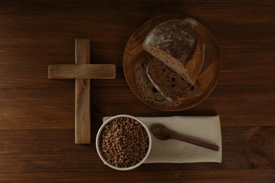 Photo of Cross, buckwheat porridge, spoon and bread on wooden table, flat lay. Lent season