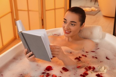 Photo of Happy woman reading book while taking bath in tub with foam and rose petals indoors