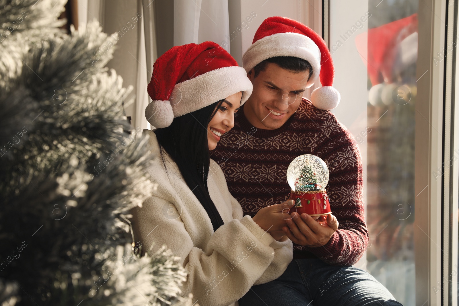 Photo of Couple in Santa hats with snow globe near window