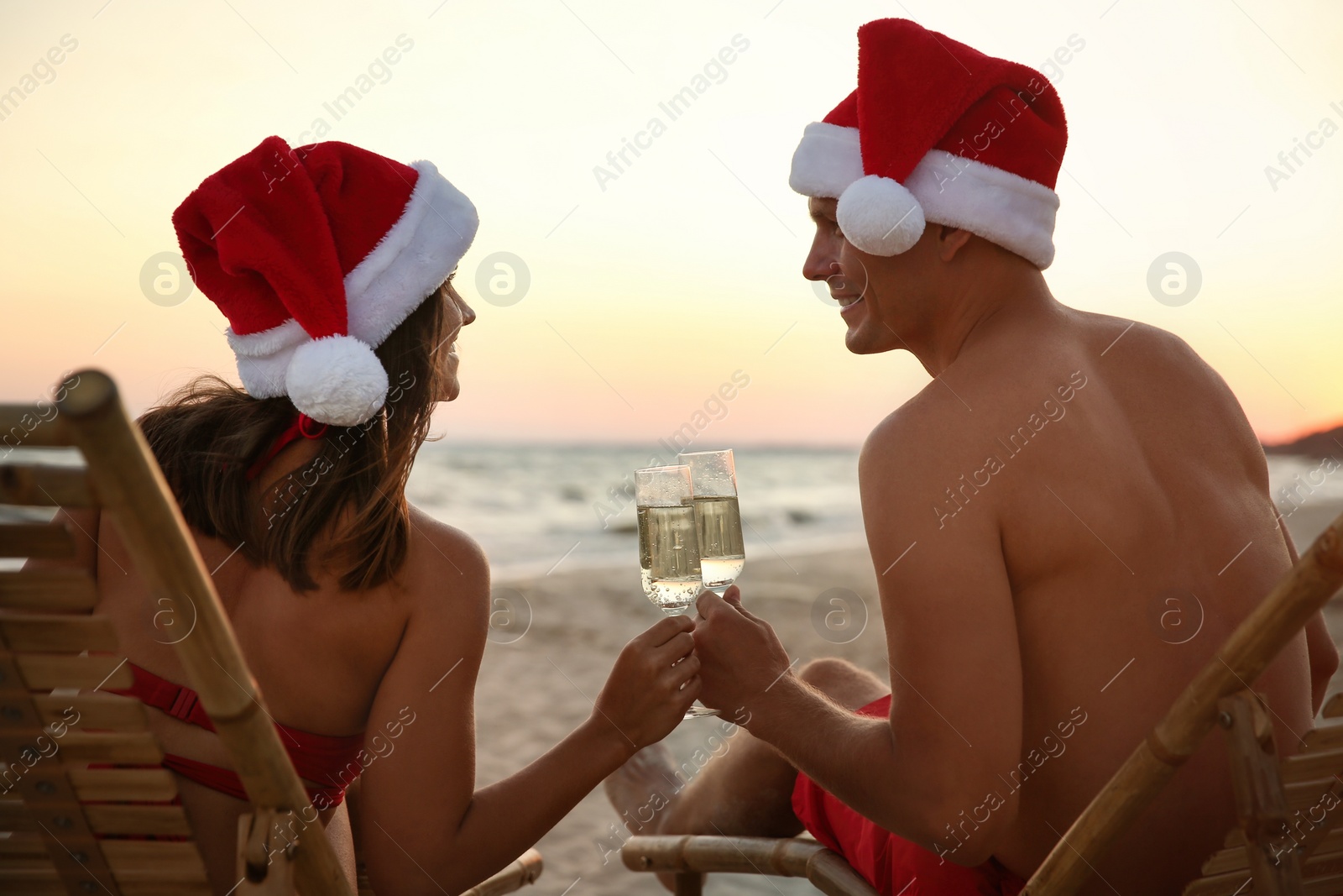 Photo of Happy couple wearing Santa hats and drinking champagne together on beach. Christmas vacation