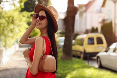 Photo of Beautiful young woman with stylish bag in red dress and sunglasses outdoors, space for text
