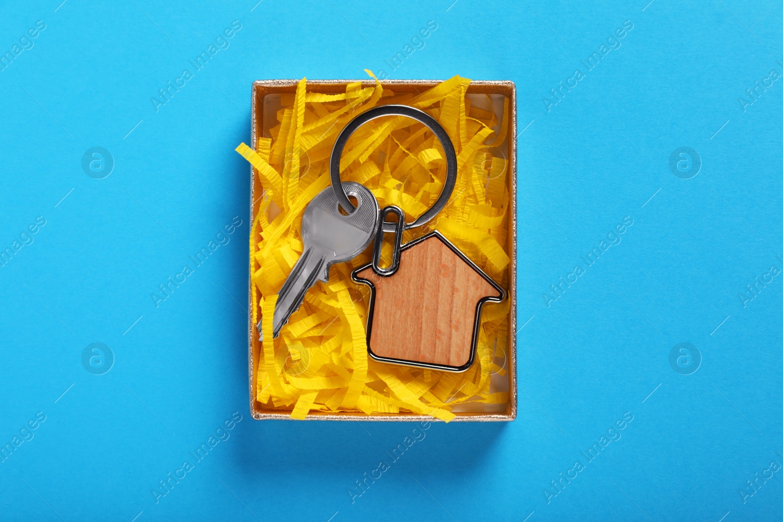 Photo of Key with trinket in shape of house and gift box on light blue background, top view. Housewarming party