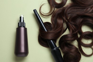 Spray bottle with thermal protection, lock of brown hair and stylish curling iron on pale green background, flat lay