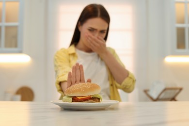 Photo of Young woman suffering from nausea at table in kitchen, focus on burger
