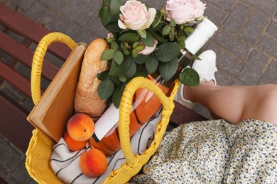 Woman sitting on bench near yellow wicker bag with roses, wine, peaches and baguette outdoors, above view