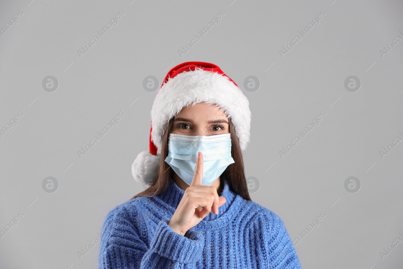 Photo of Pretty woman in Santa hat and medical mask showing silence gesture on grey background