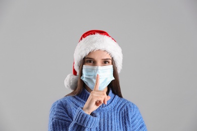 Photo of Pretty woman in Santa hat and medical mask showing silence gesture on grey background