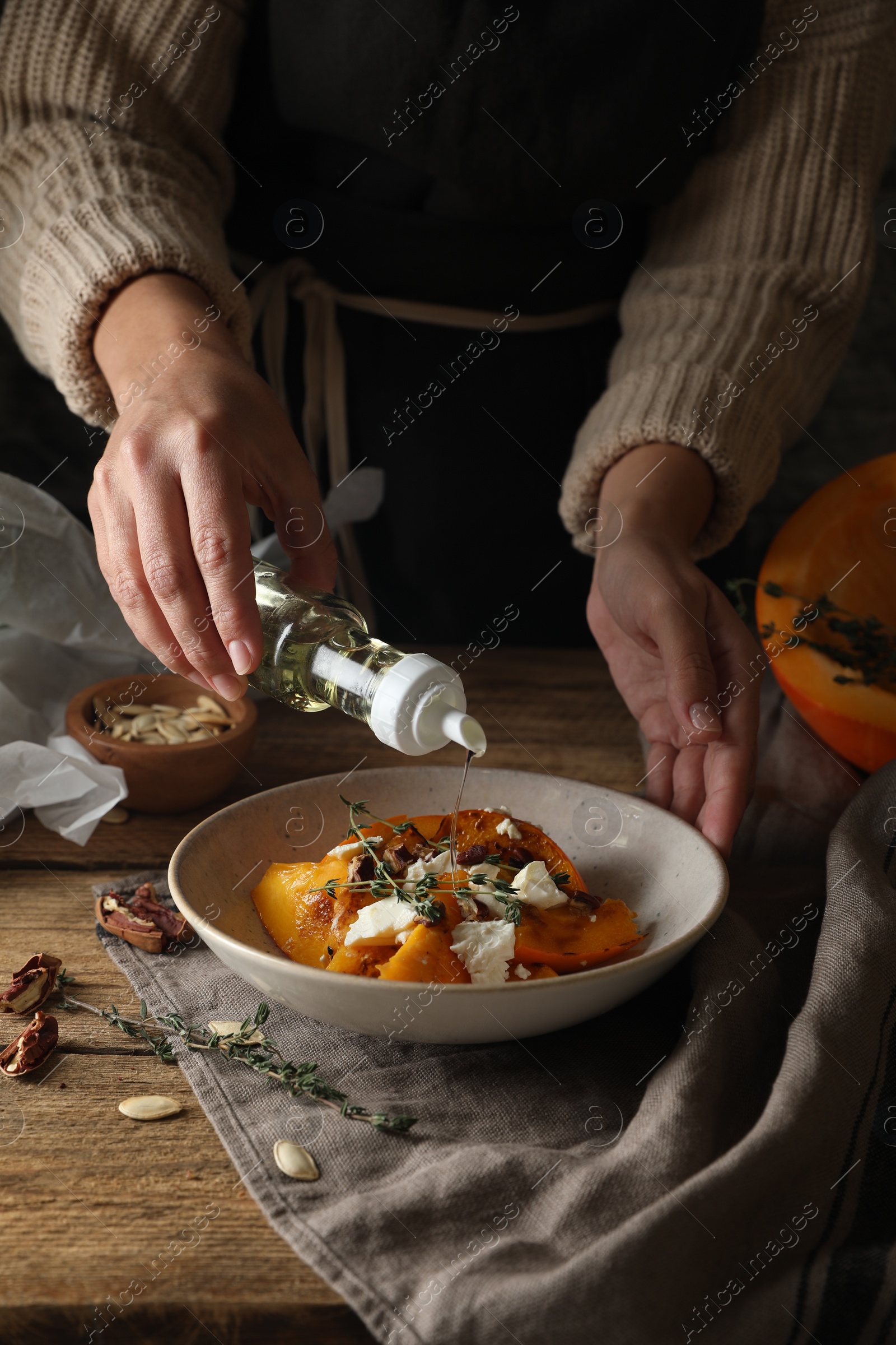 Photo of Woman adding cooking oil to freshly baked pumpkin slices with cheese, thyme and pecans at wooden table, closeup