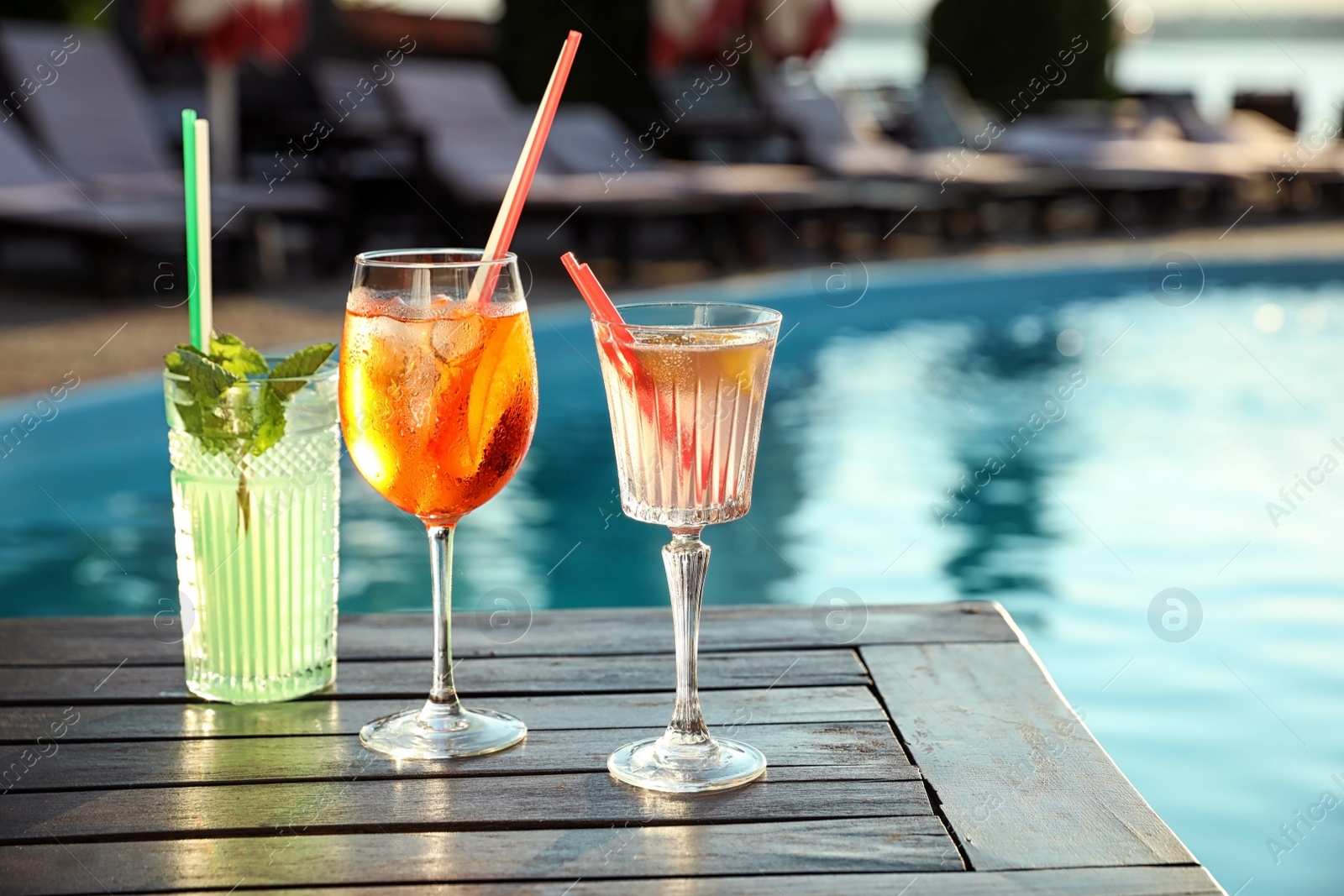 Photo of Glasses of fresh summer cocktails on wooden table near swimming pool outdoors