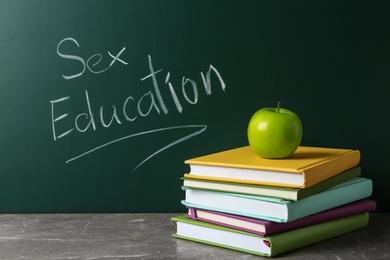 Photo of Books and apple on grey table near chalkboard with phrase "Sex education"