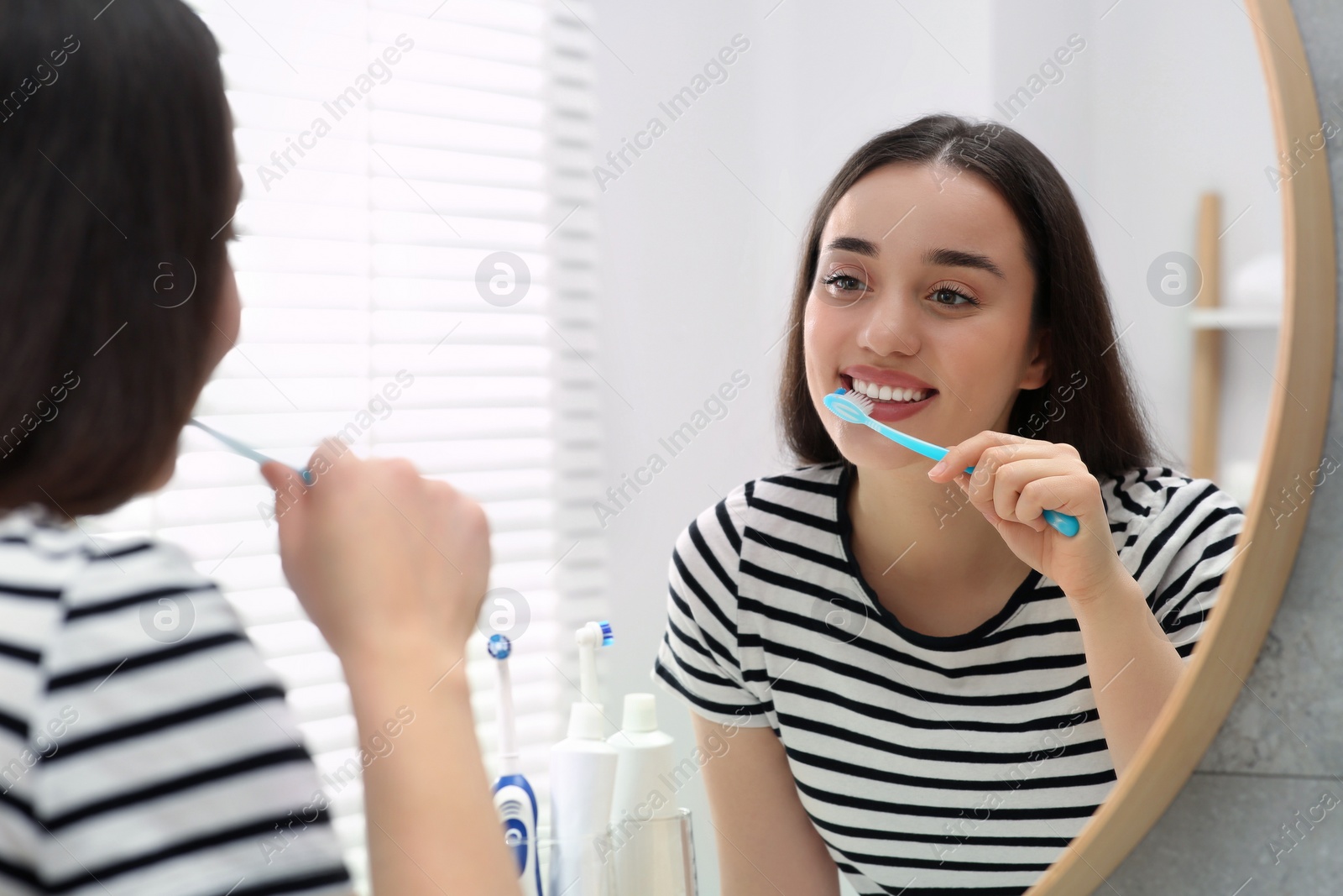 Photo of Young woman brushing her teeth with plastic toothbrush near mirror in bathroom