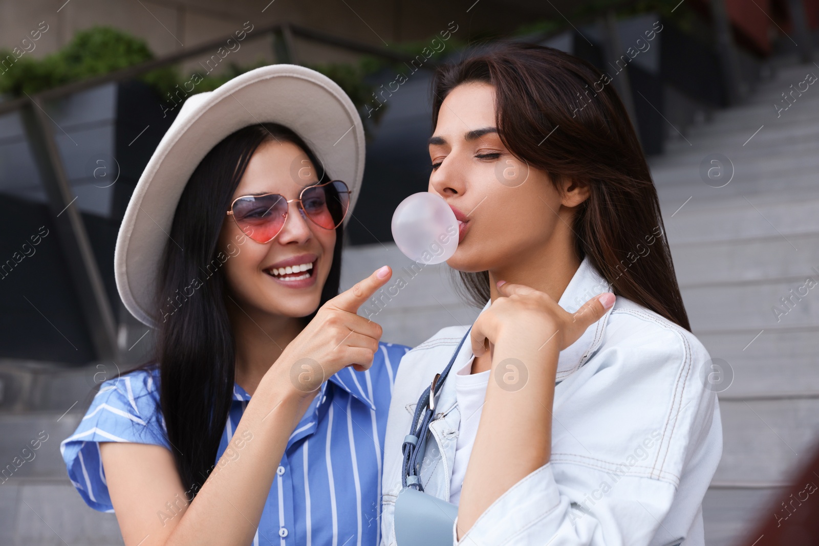 Photo of Stylish women having fun and blowing gum near stairs outdoors