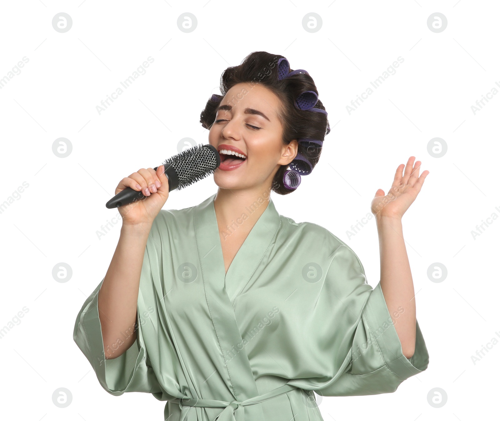 Photo of Happy young woman in silk bathrobe with hair curlers singing into hairbrush on white background