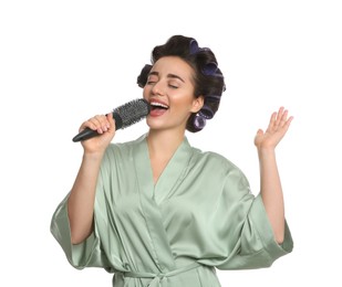 Happy young woman in silk bathrobe with hair curlers singing into hairbrush on white background