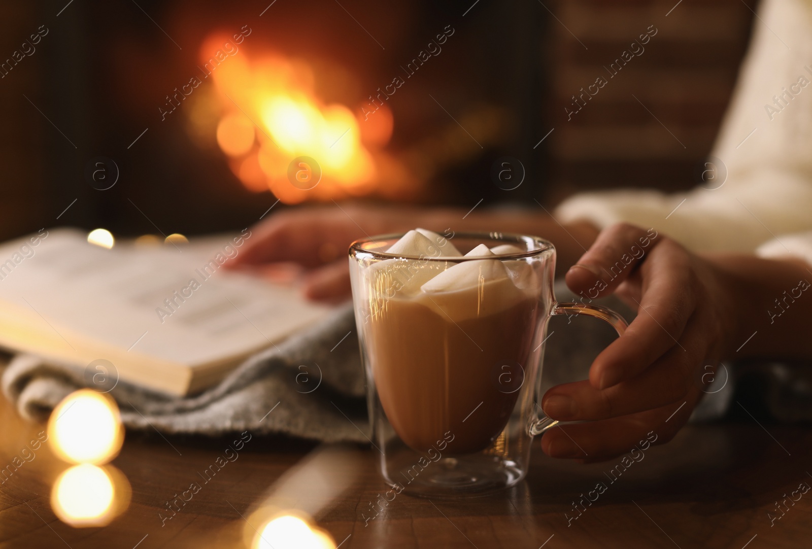 Photo of Woman with cocoa and book near burning fireplace indoors, closeup