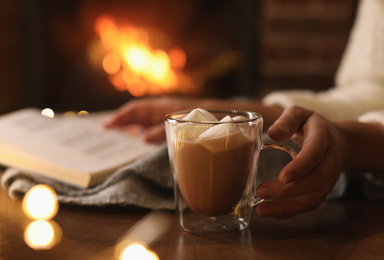Photo of Woman with cocoa and book near burning fireplace indoors, closeup