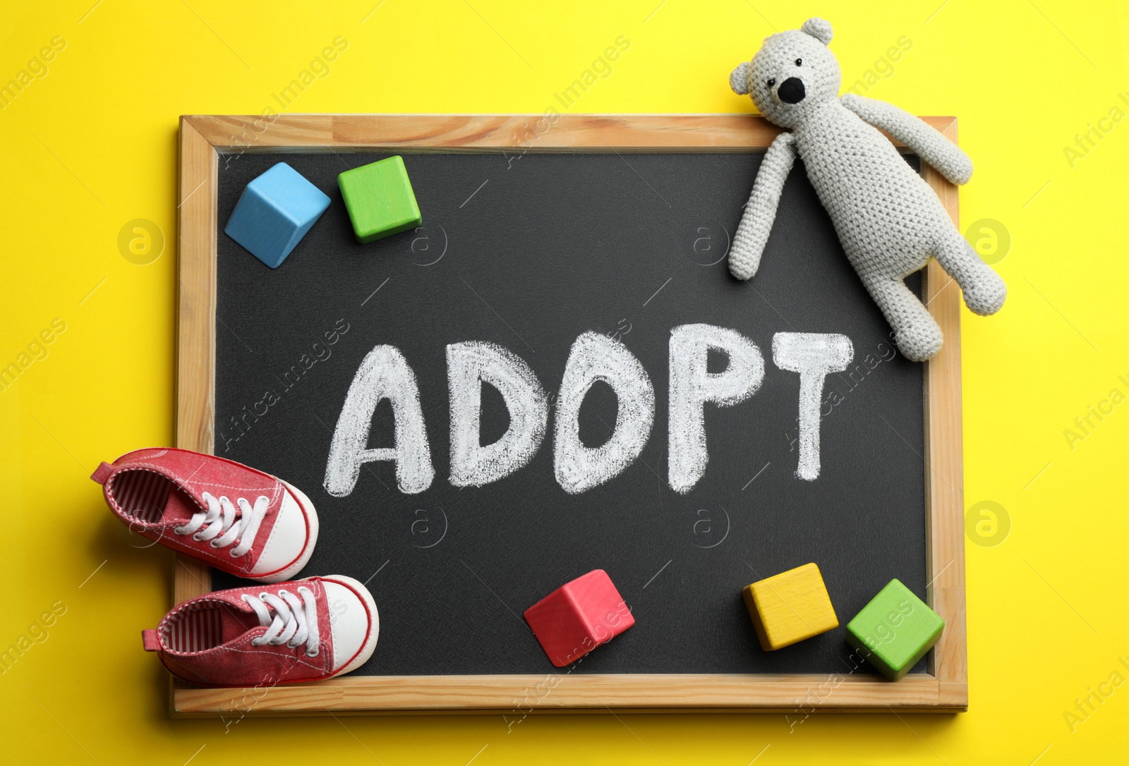 Photo of Small chalkboard with word ADOPT, toy bear, colorful cubes and baby shoes on yellow background, top view