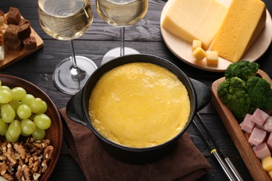 Photo of Fondue pot with melted cheese, glasses of wine and different products on black wooden table, above view