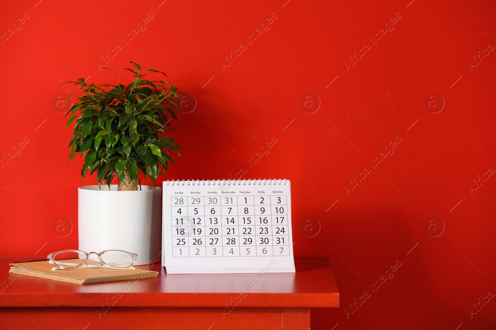 Photo of Paper calendar and plant on table near red wall. Space for text