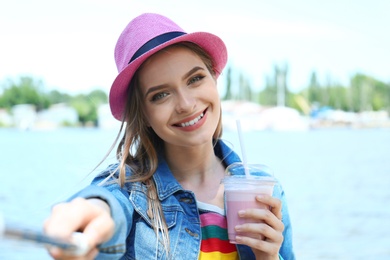 Photo of Happy young woman with drink taking selfie on riverside