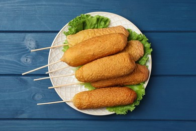 Photo of Delicious deep fried corn dogs and lettuce leaves on blue wooden table, top view