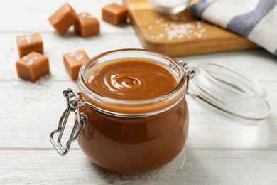 Photo of Yummy salted caramel in glass jar and candies on white wooden table