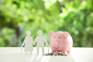 Photo of Pension savings. Figure of senior couple, piggy bank and coins on white table against blurred green background