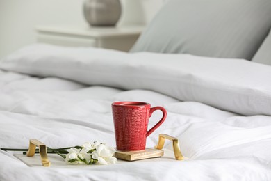Photo of Mug of hot drink with stylish cup coaster and beautiful freesia flowers on bed in room