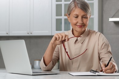 Photo of Beautiful senior woman taking notes near laptop at white marble table in kitchen