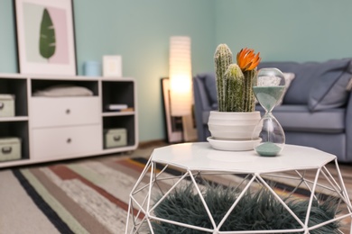 Photo of Stylish table with hourglass and cactus in living room