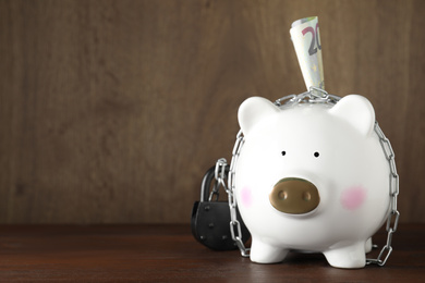 Photo of Piggy bank with steel chain, padlock and banknote on wooden table, space for text. Money safety concept