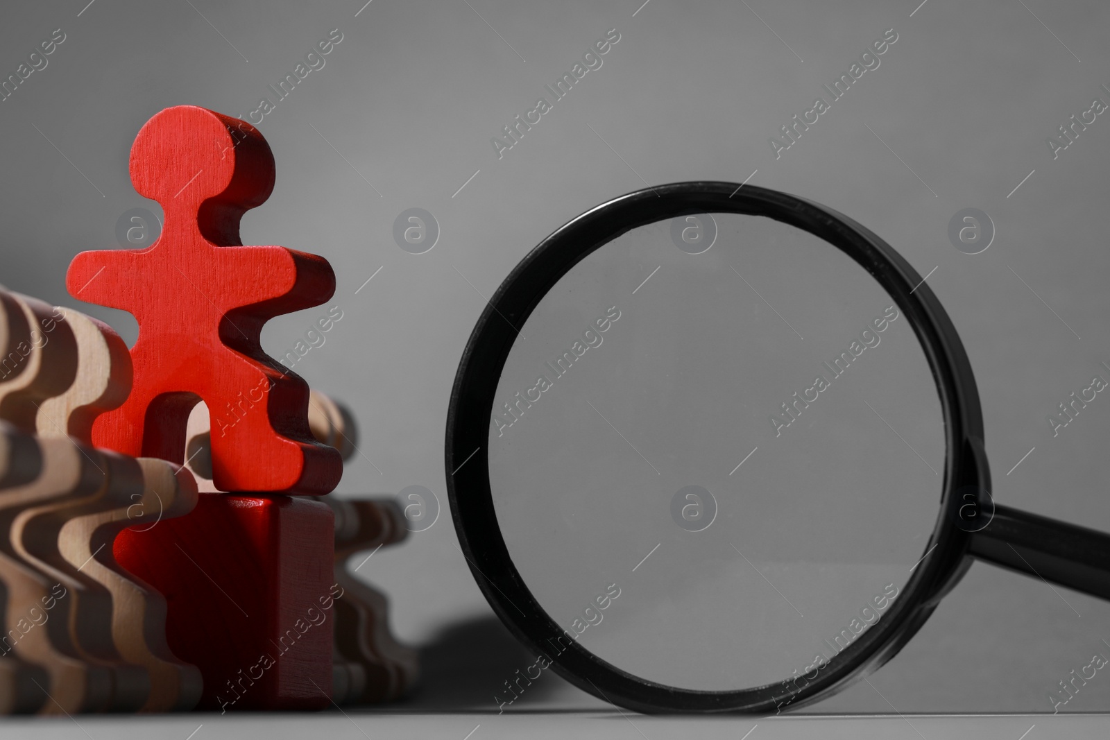 Photo of Red figure among wooden ones near magnifying glass on grey background, closeup. Recruiter searching employee