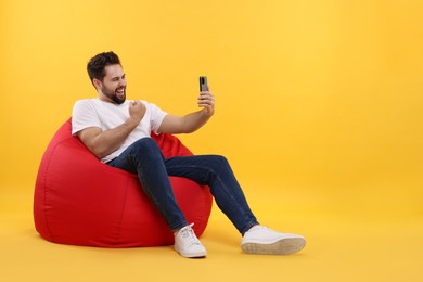 Photo of Happy young man using smartphone on bean bag chair against yellow background, space for text