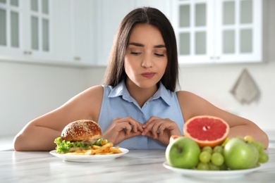 Photo of Woman choosing between fruits and burger with French fries in kitchen