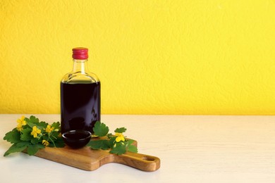Photo of Bottle of celandine tincture and plant on white table against yellow background, space or text