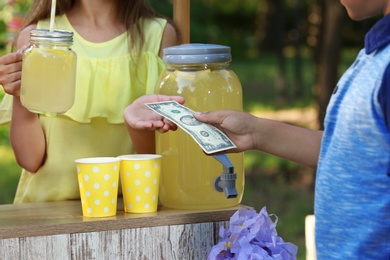 Photo of Little girl selling natural lemonade to African-American boy in park, closeup. Summer refreshing drink