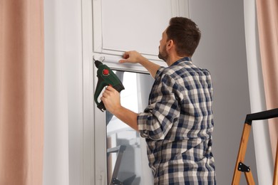 Photo of Man with drill installing roller window blind on stepladder indoors