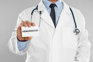Photo of Doctor holding business card with word HEMORRHOID on light grey background, closeup