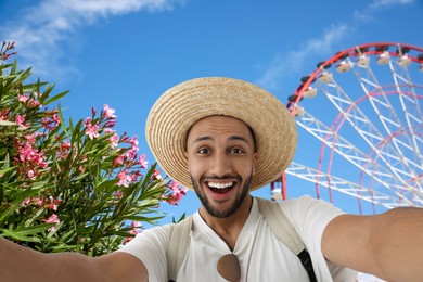 Image of Smiling young man in straw hat taking selfie near observation wheel