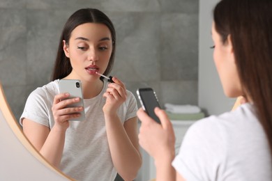 Photo of Beautiful young woman using smartphone while applying lipstick near mirror indoors. Internet addiction