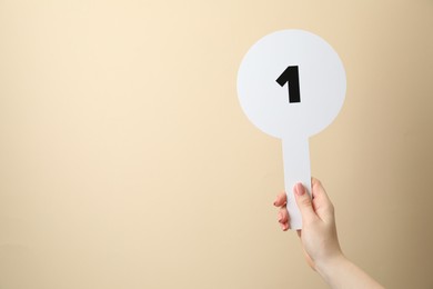 Woman holding auction paddle with number 1 on beige background, closeup. Space for text