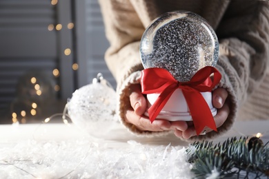 Photo of Woman holding Christmas snow globe with red bow on blurred background, closeup. Space for text