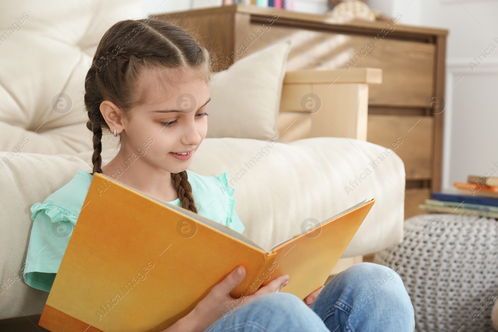 Photo of Cute little girl reading book near sofa at home