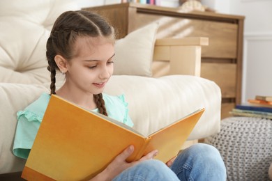 Photo of Cute little girl reading book near sofa at home