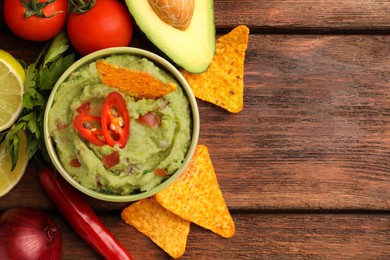 Photo of Bowl of delicious guacamole, nachos chips and ingredients on wooden table, flat lay. Space for text