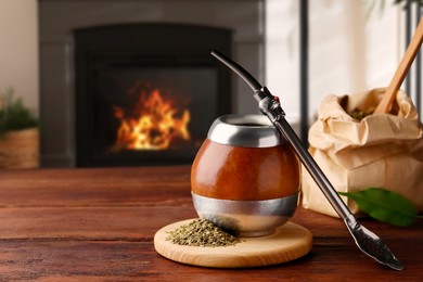 Image of Calabash with mate tea and bombilla on wooden table near fireplace. Space for text