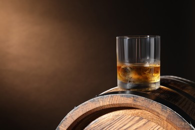Photo of Glass of tasty whiskey on wooden barrel against dark background, space for text