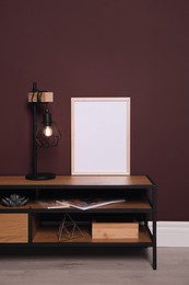 Photo of Empty frame and stylish lamp on wooden table near brown wall. Mockup for design